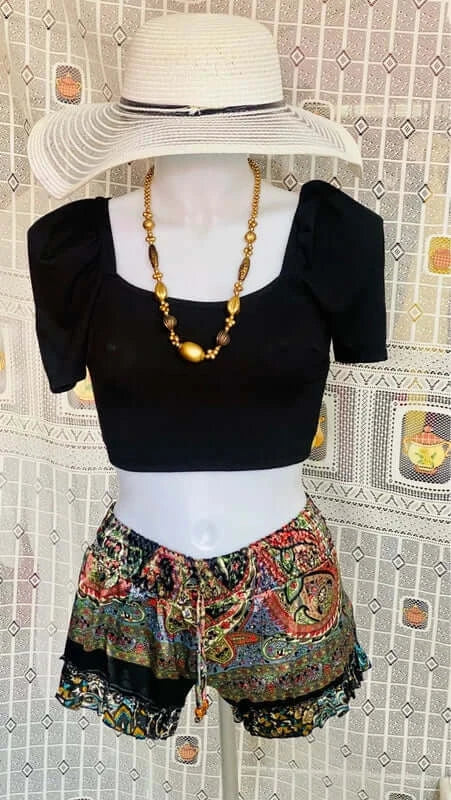 Stylish boho chic outfit featuring a black puff sleeve top and colorful patterned shorts with a straw hat and necklace.